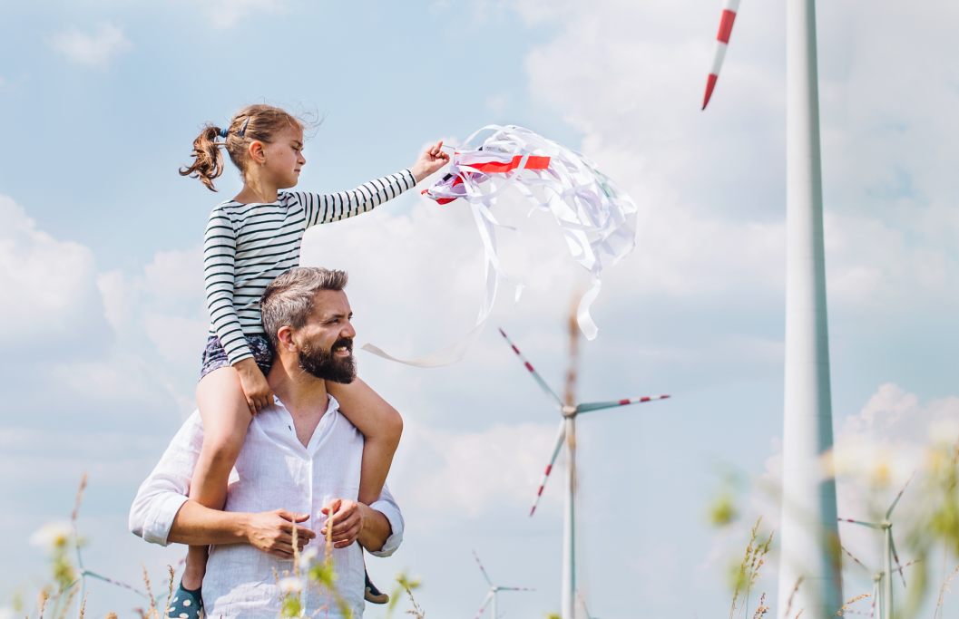 Vater trägt Tochter auf den Schultern auf einem Feld mit Windrädern im Hintergrund unter blauem Himmel mit Wolken.