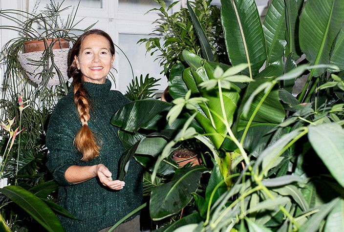 Eine Frau steht hinter ein paar 2 Meter großen Zimmerpflanzen in ihrer Wohnung und lächelt.